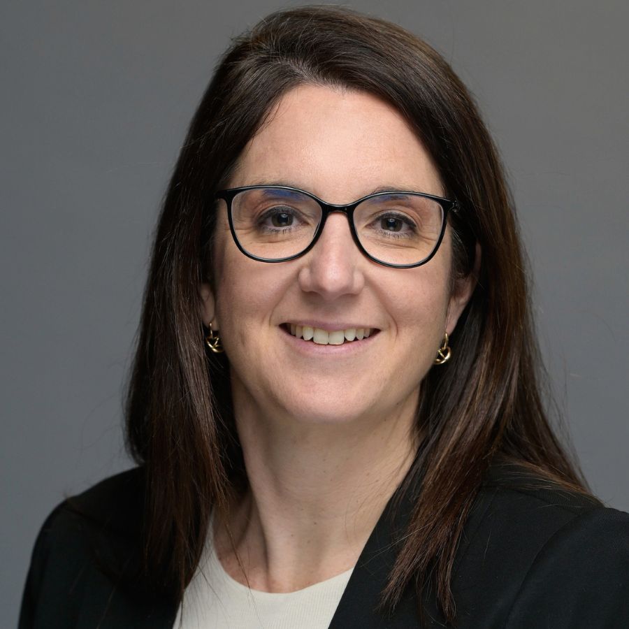A woman with long brown hair and glasses, wearing a black blazer and a light-colored top, smiles at the camera against a plain gray background.