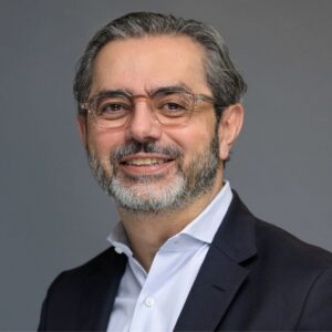 A middle-aged man with glasses, gray hair, and a beard, wearing a dark suit jacket over a light-colored shirt, poses against a plain gray background.