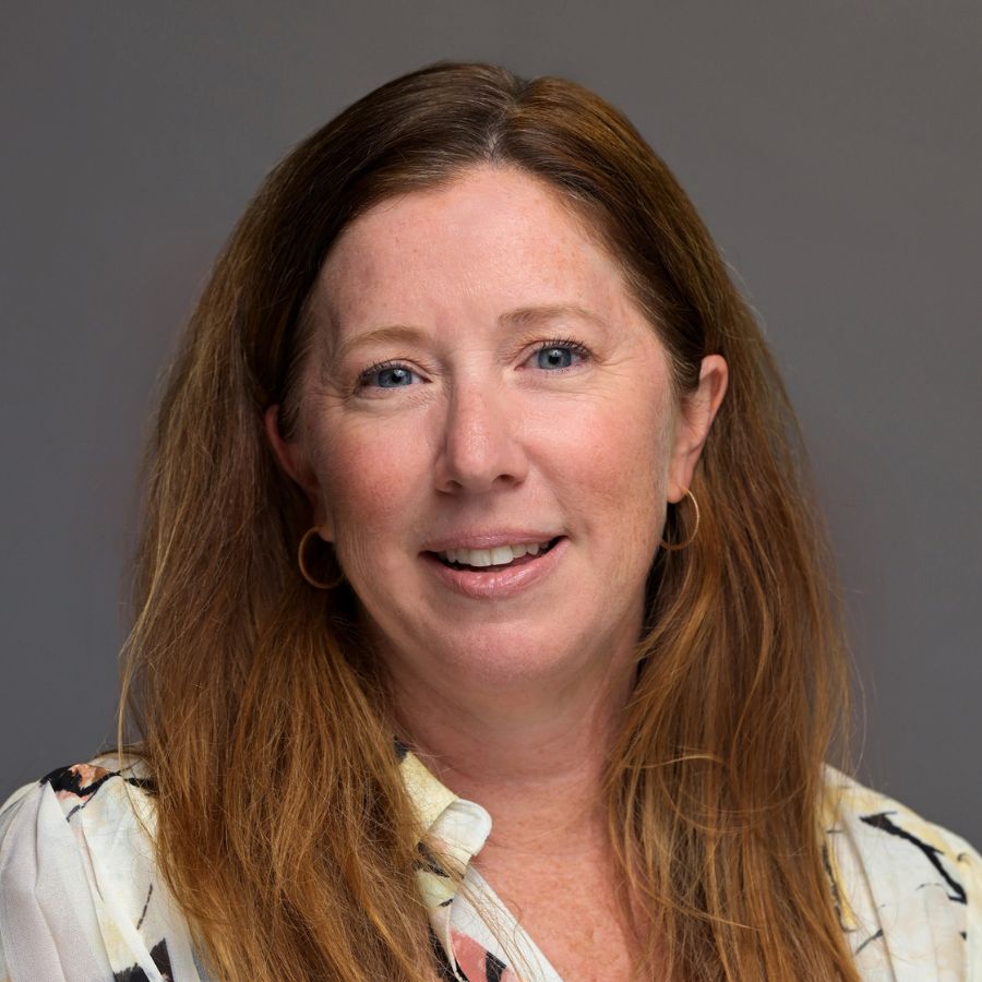 A woman with long reddish-brown hair, wearing a patterned blouse and hoop earrings, smiles in front of a plain gray background.